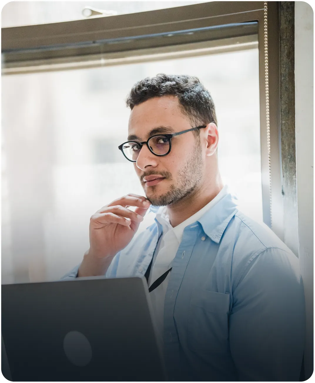 Man in glasses and a light blue shirt holding a laptop by a bright window, resting a hand on his chin