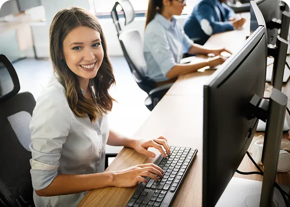Smiling woman typing at a keyboard at her desk in an open office as colleagues work at computers behind her