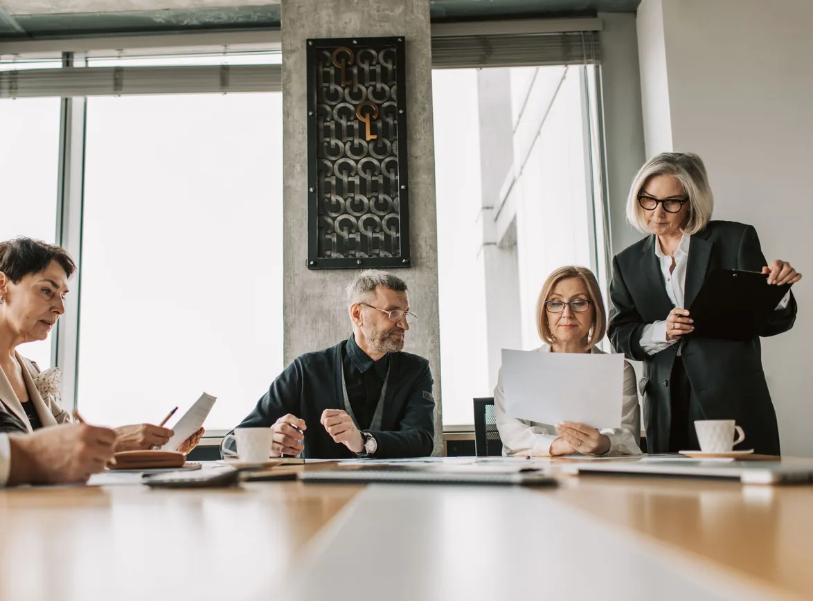 Senior executives review papers around a boardroom table while a colleague stands holding a clipboard