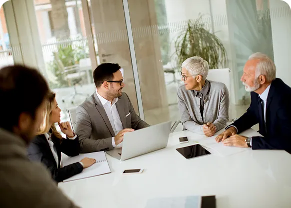 Business meeting at a white conference table, a man at a laptop talking with older colleagues as others listen