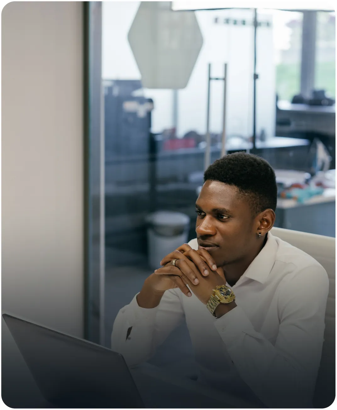 Man in a white shirt seated at a laptop in an office, hands clasped under his chin
