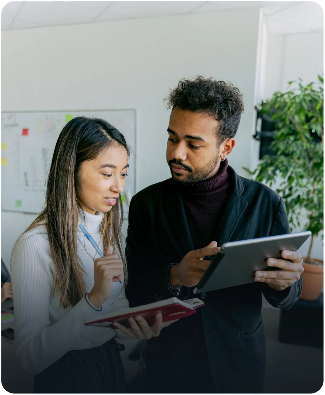 Two colleagues standing in an office reviewing a tablet and notebook in front of a whiteboard with sticky notes