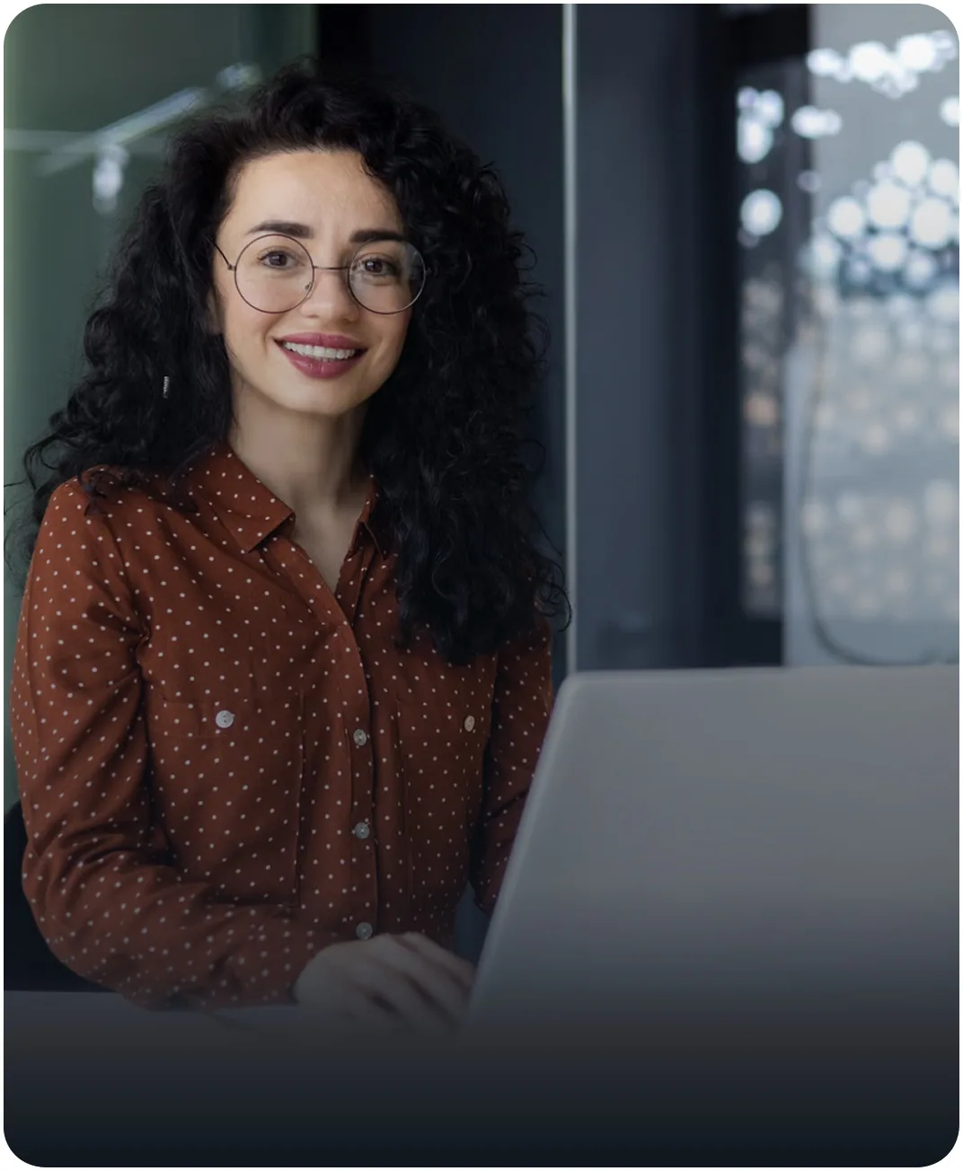 Smiling professional with round glasses and curly hair working on a laptop in a modern office