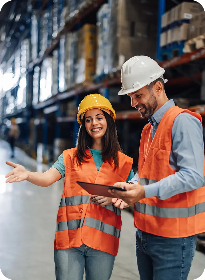 Two warehouse workers in hard hats and safety vests reviewing a tablet between tall storage racks