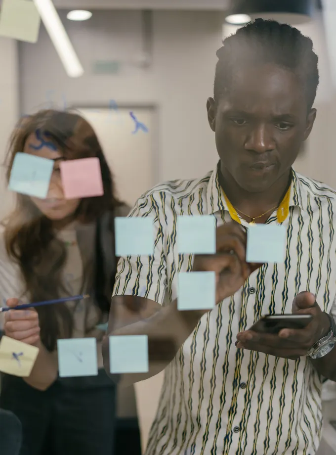 Marketing team member checking a phone while arranging sticky notes on a glass wall as a colleague works nearby