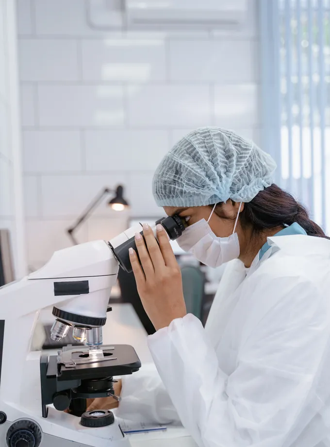 Scientist in a hairnet, face mask and white coverall looking through a microscope in a pharmaceutical lab