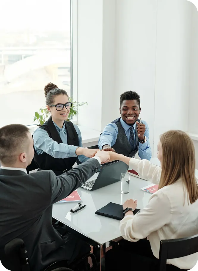 Four business consultants bumping fists over a meeting table with a laptop and notebooks in a bright office