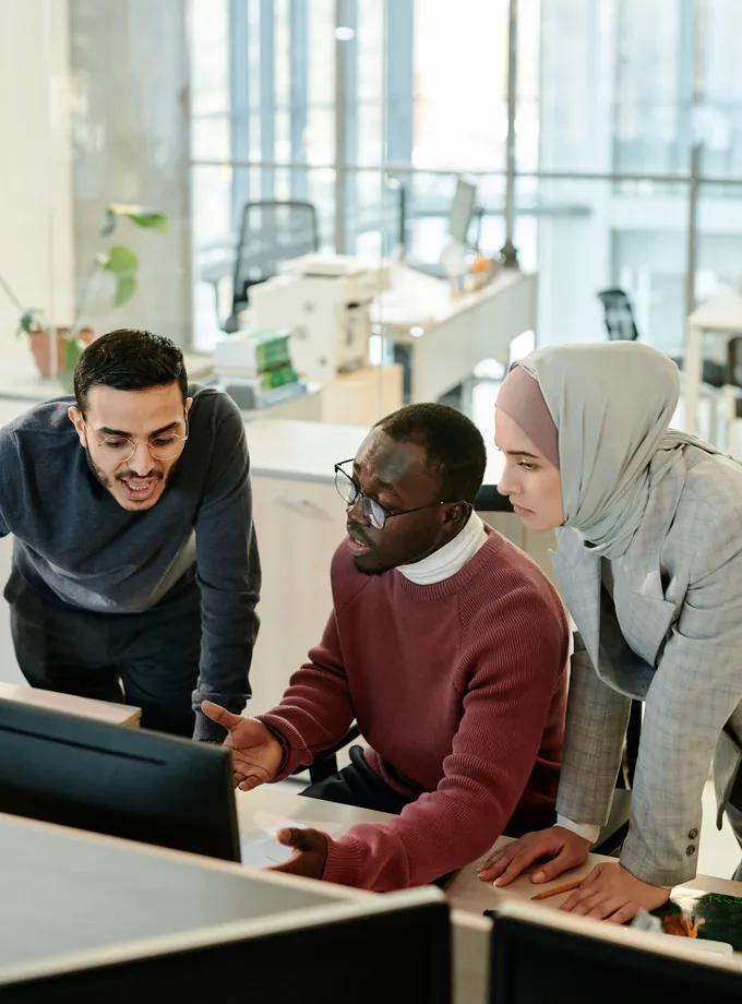 Three colleagues lean over a computer monitor talking through a task in a bright open-plan tech office