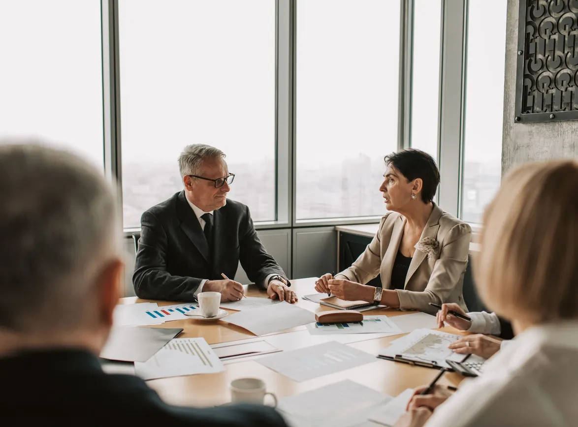 Senior executives in a boardroom meeting at a table covered with charts beside floor-to-ceiling windows