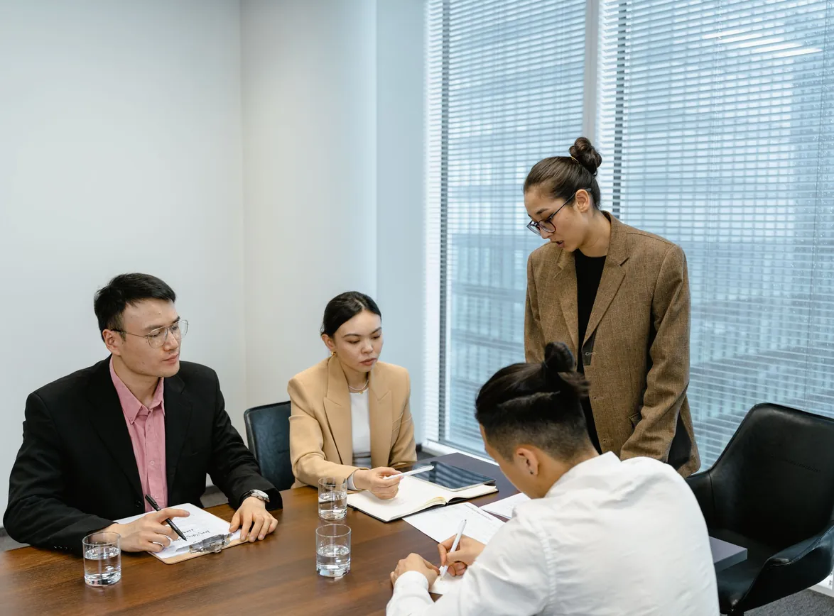 Project lead standing at a conference table guiding three seated colleagues as they take notes