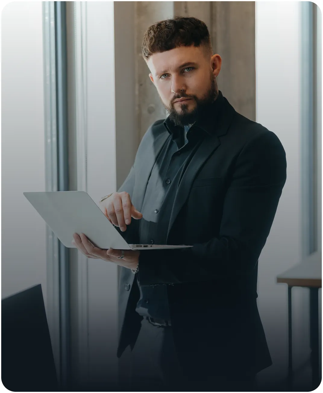 Bearded professional in a dark suit holding an open laptop while standing by an office window