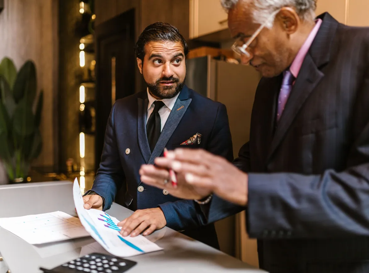 Two businessmen in suits reviewing printed bar charts at a desk with a calculator