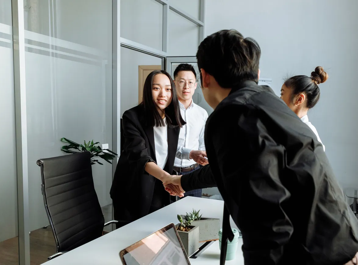 Woman in a black blazer shaking hands with a man across a desk as colleagues look on in a glass-walled office