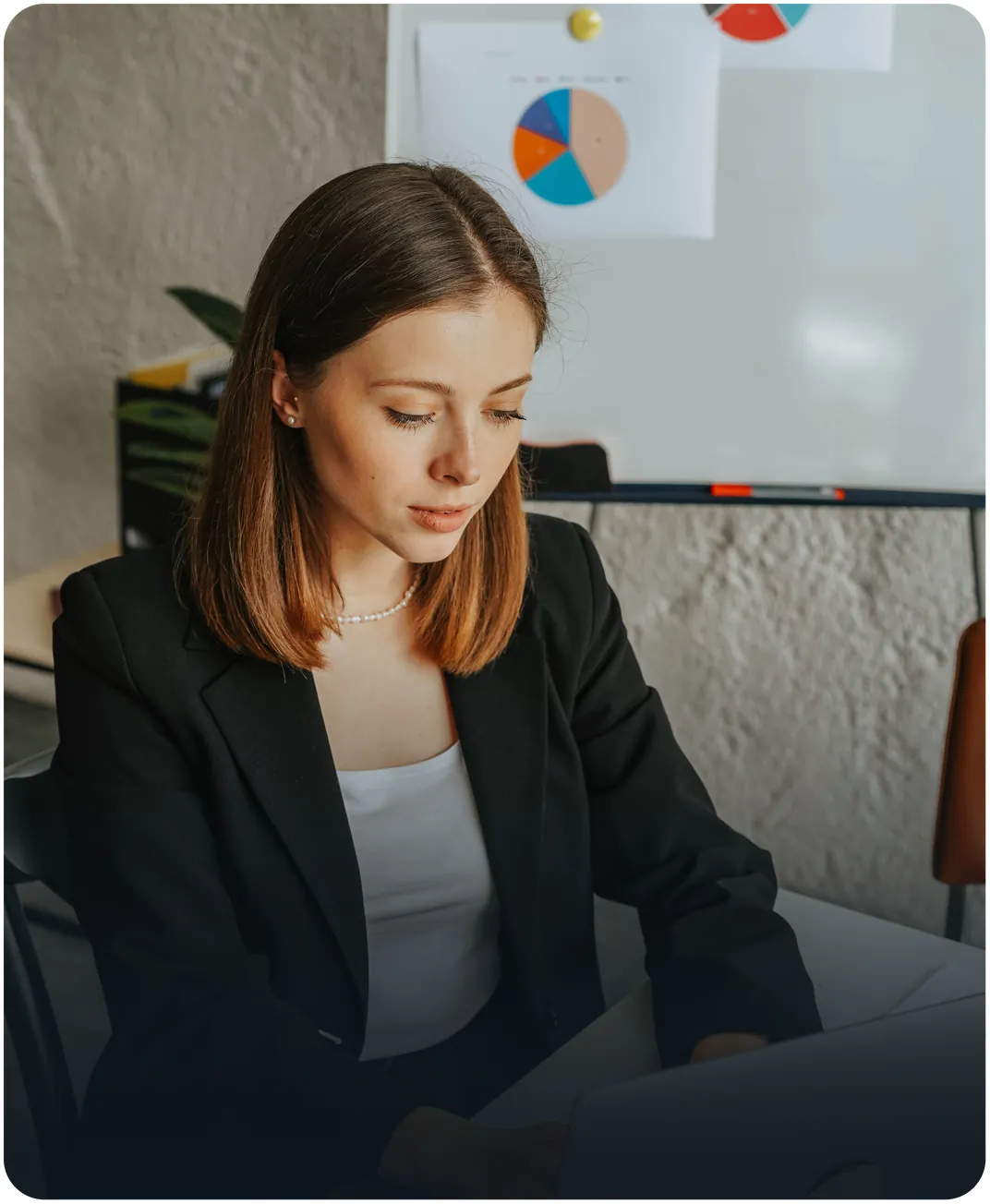 Businesswoman in a black blazer working at her desk with pie chart printouts pinned to a whiteboard behind her