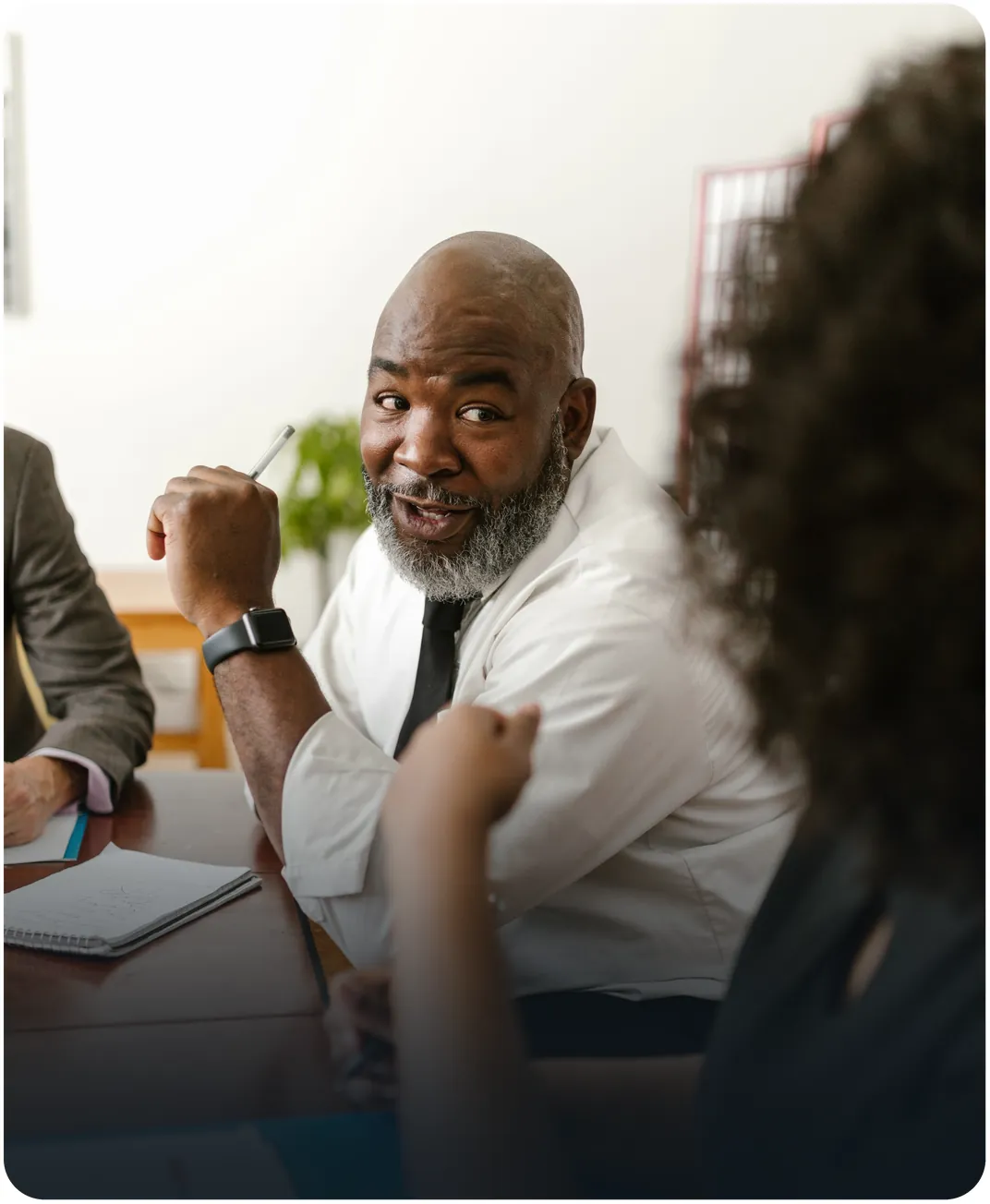Bearded businessman holding a pen and talking with colleagues around a conference table with an open notebook