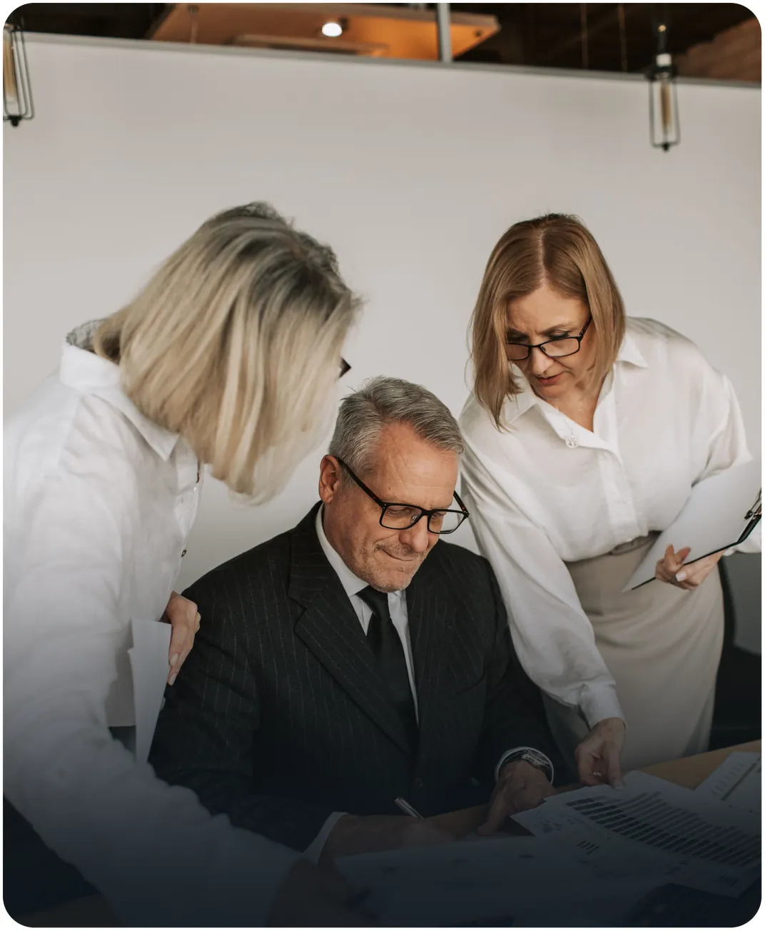 Two colleagues lean over a senior executive at his desk as he reviews printed bar chart reports