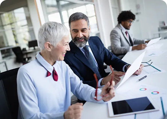 Gray-haired businesswoman and a bearded colleague in a suit smiling over printed charts at an office table