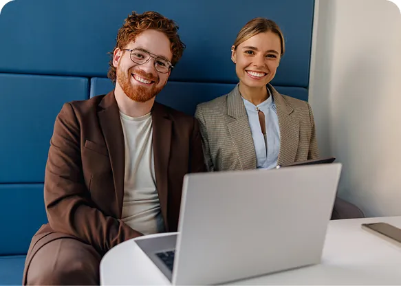 Two smiling colleagues in blazers sitting side by side on a blue booth bench behind a laptop