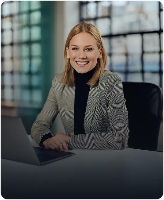 Smiling businesswoman in a houndstooth blazer and black turtleneck seated at a laptop in an office