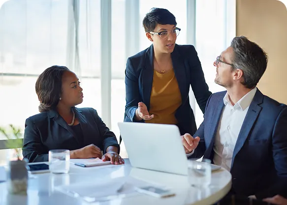 Woman in glasses standing and explaining a point to two seated colleagues at a round table with a laptop