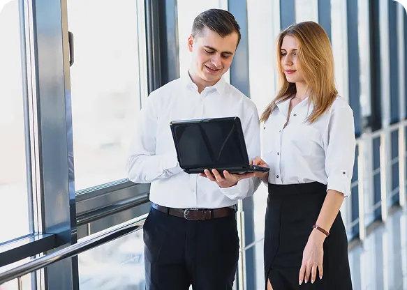 Two colleagues in white shirts looking at a laptop together in a bright office corridor with tall windows