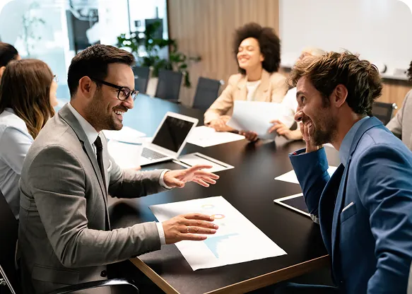 Two men laughing over printed charts during a team meeting at a conference table with colleagues and a laptop