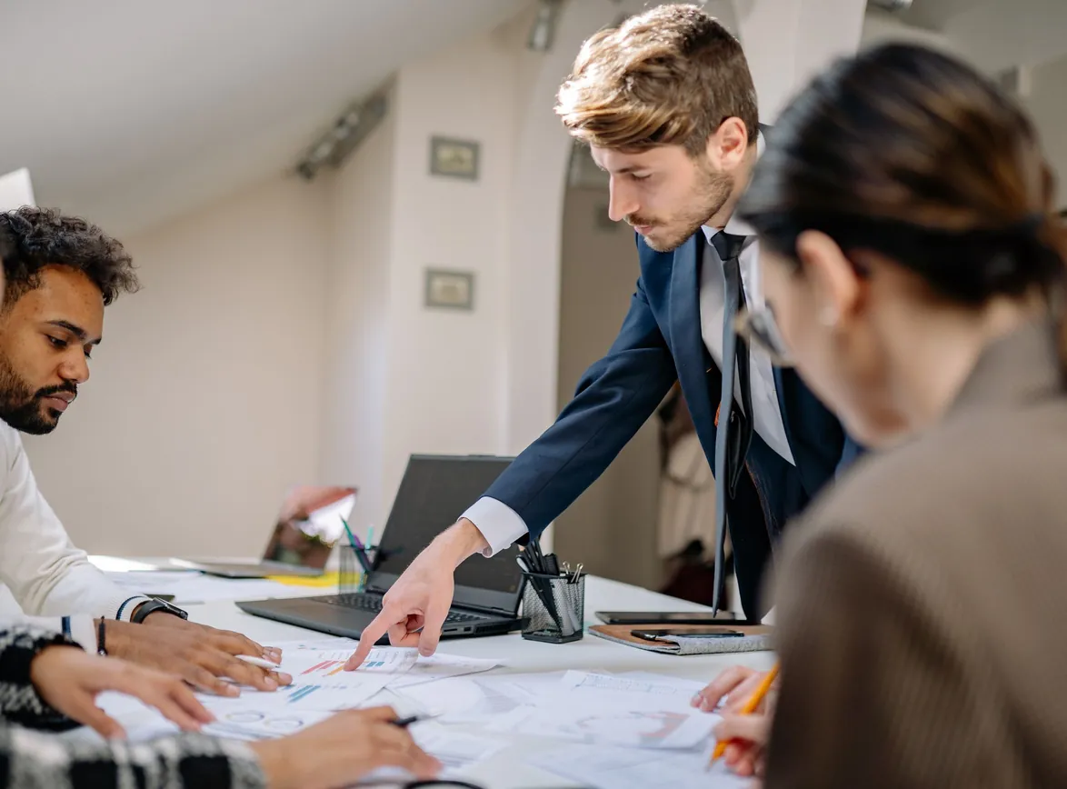 Manager in a suit leaning over a table and pointing to printed charts as team members take notes