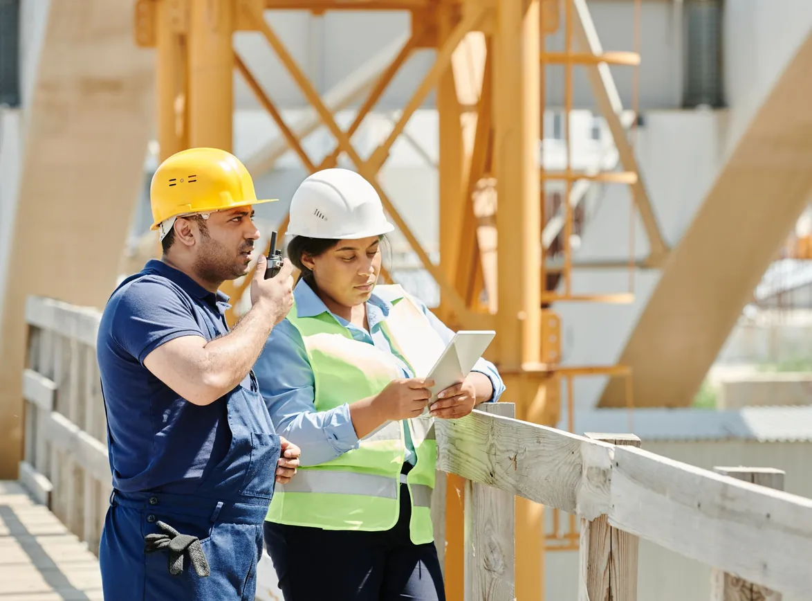 Construction worker on a walkie-talkie beside a colleague in a hi-vis vest reading a tablet on a building site