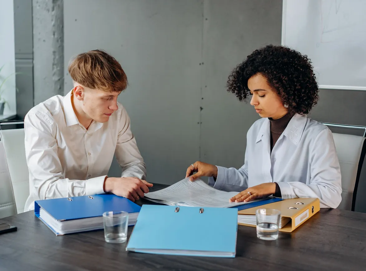 Two colleagues reviewing printed documents from ring binders at an office conference table
