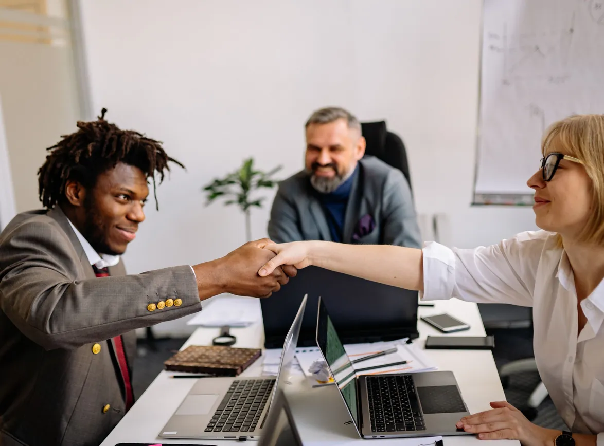 Two professionals shaking hands across a desk with laptops while a smiling colleague looks on in an office