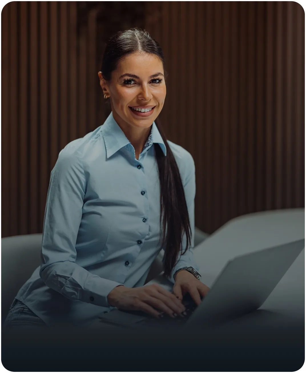 Smiling businesswoman in a light blue shirt typing on a laptop in front of a wood slat wall