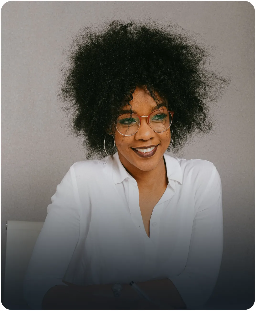 Smiling businesswoman in round glasses and a white blouse seated with her arms resting on a desk