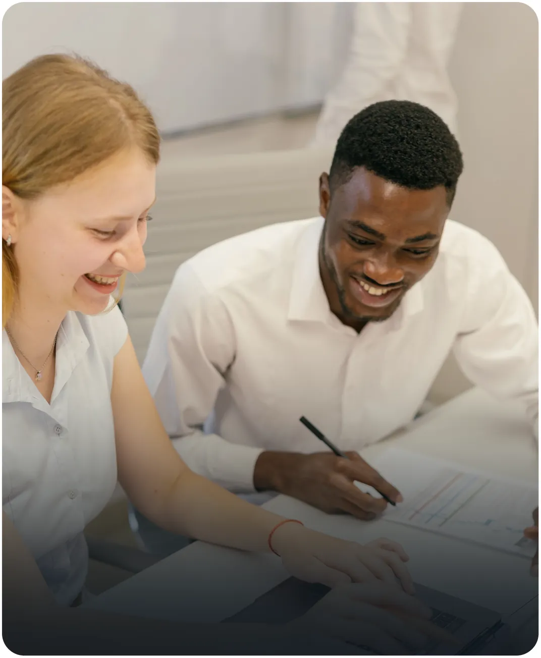 Two smiling colleagues at a desk, one marking up a printed chart with a pen while the other uses a laptop
