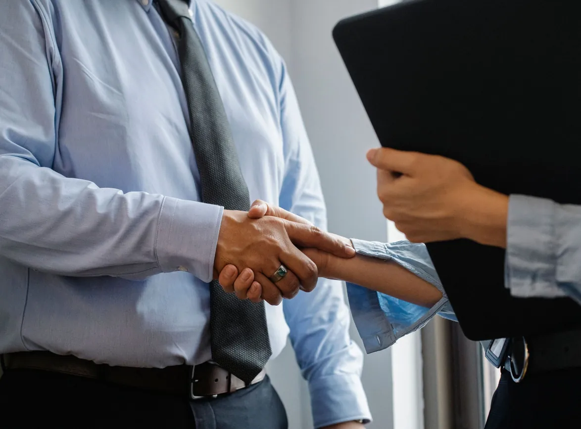 Man in formal shirt and tie shaking hand of woman with black folder while meeting in office