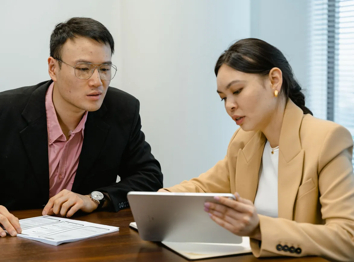 Two colleagues at an office desk reviewing information on a tablet alongside a printed form