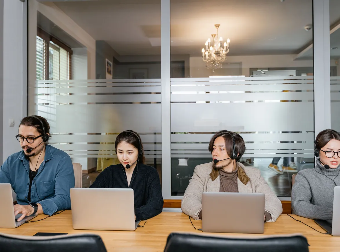 Four sales reps wearing headsets working on laptops side by side at a long office desk