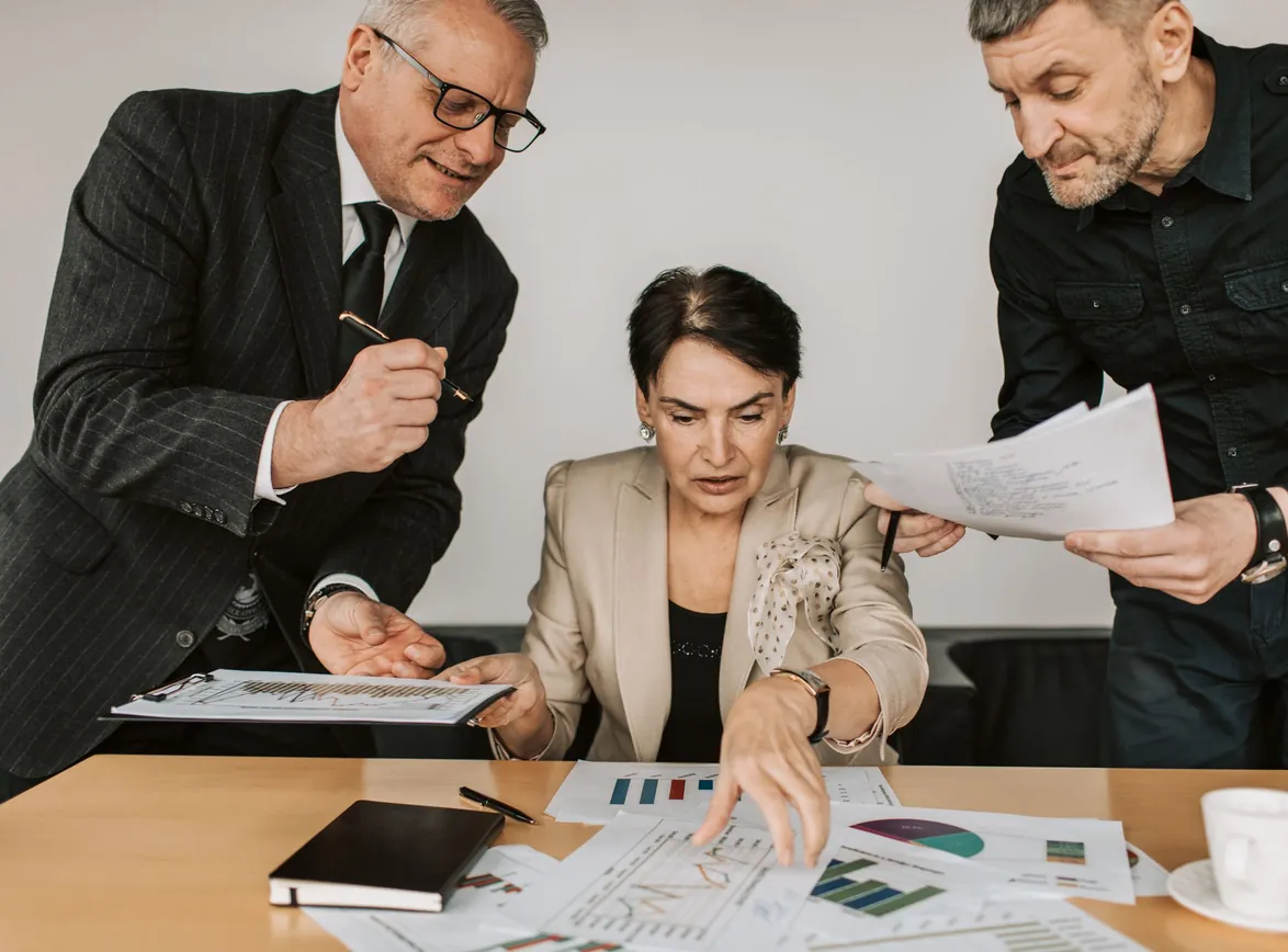 Executive pointing to printed charts on a table as two colleagues review reports beside her