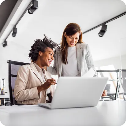Two businesswomen smiling as they review a laptop together at a desk in a bright office