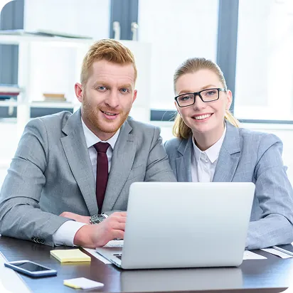 Two smiling professionals in gray suits seated together behind a laptop at an office desk