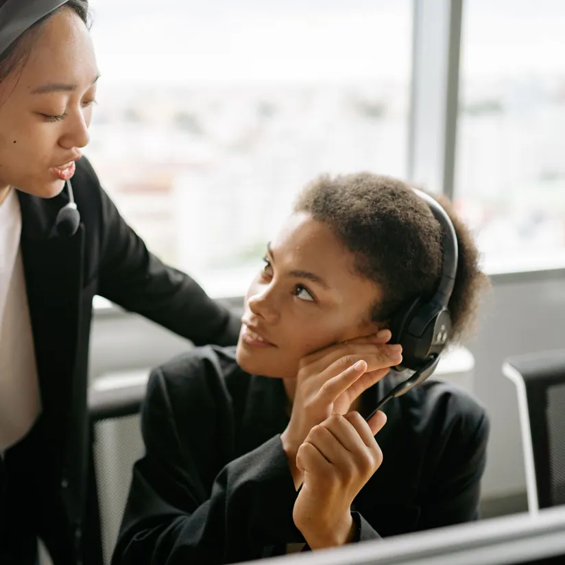 Team lead wearing a headset leans in to talk with a seated sales rep who listens through her own headset
