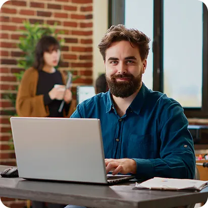 Bearded man in a teal shirt working on a laptop in a brick-walled office as a coworker uses a tablet behind him