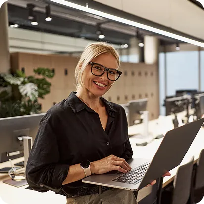 Smiling woman in glasses typing on a laptop she holds in an open office lined with desktop monitors