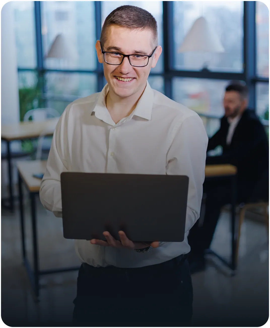 Smiling man in glasses and a white shirt holding a laptop in an office with large windows, a colleague behind