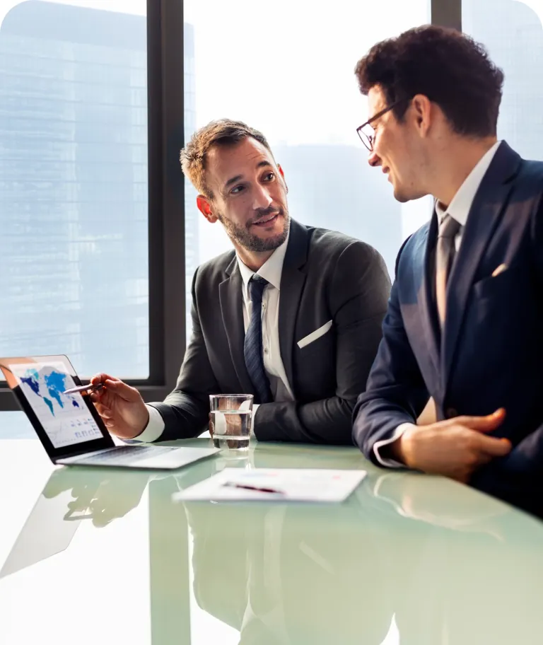 Two businessmen in suits talking at a glass conference table as one points a pen at a world map chart on a laptop