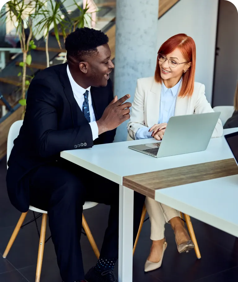 Businessman in a dark suit talking with a smiling colleague in a cream blazer who sits at a laptop