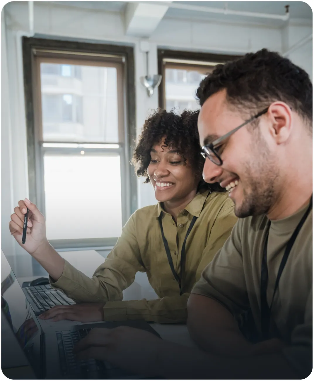 Two smiling colleagues wearing lanyards working on laptops side by side near an office window