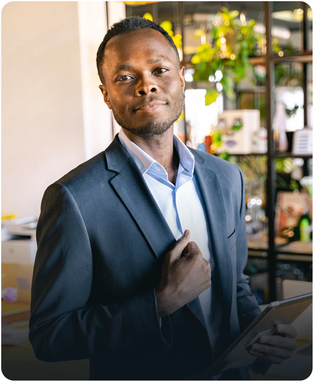 Businessman in a navy suit jacket holding a tablet in a bright open office with shelves and plants