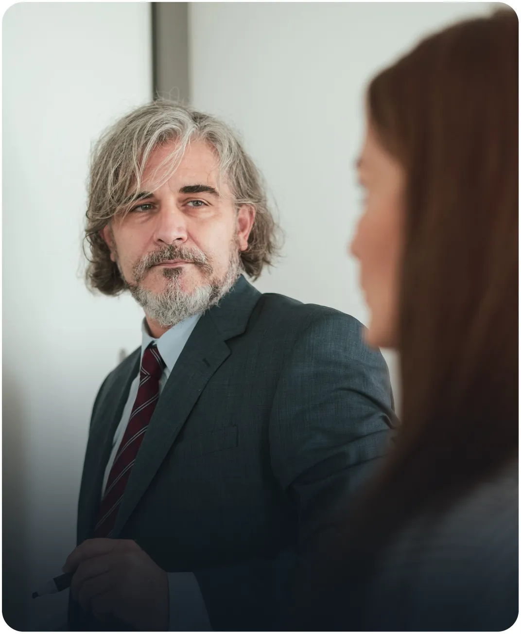 Gray-haired businessman in a suit and tie holding a marker as he listens to a colleague in a meeting