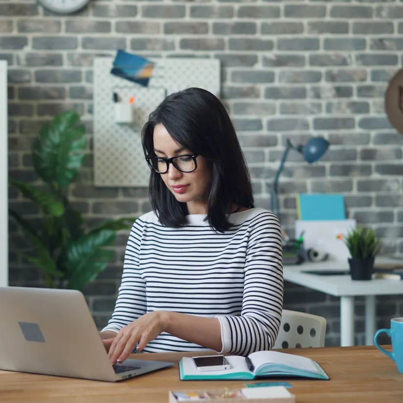 Professional typing on a laptop at a wooden desk in a bright office with a brick wall