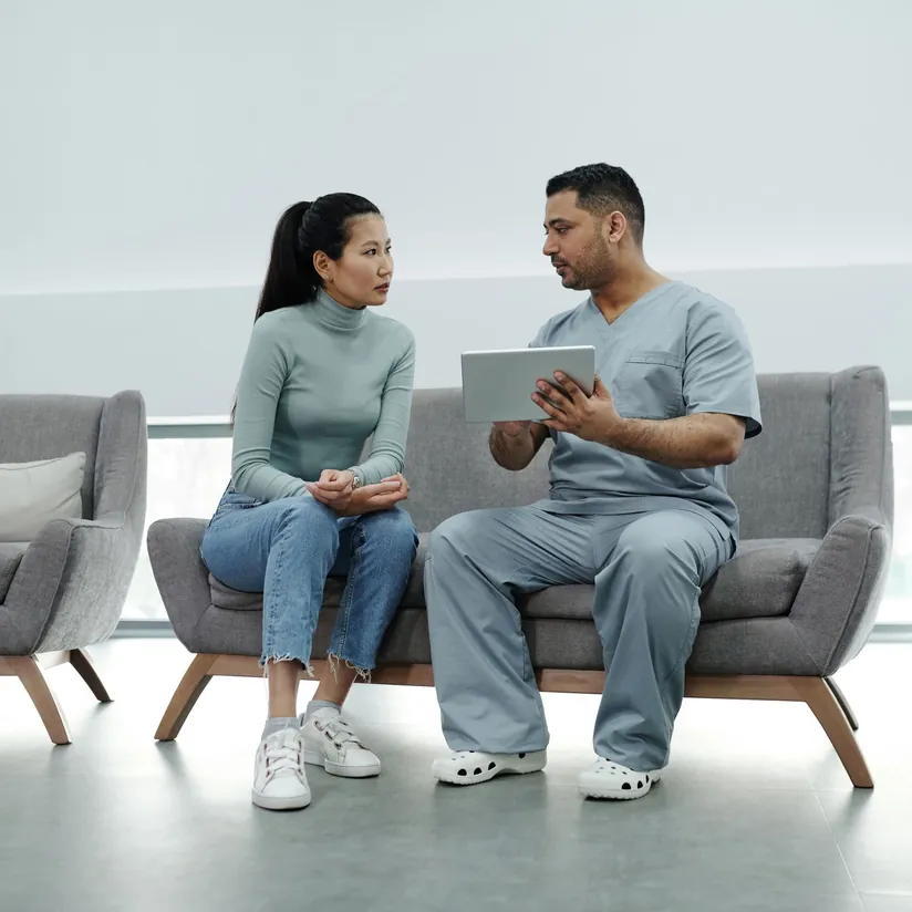 Clinician in scrubs showing a tablet to a patient seated beside him on a waiting room sofa